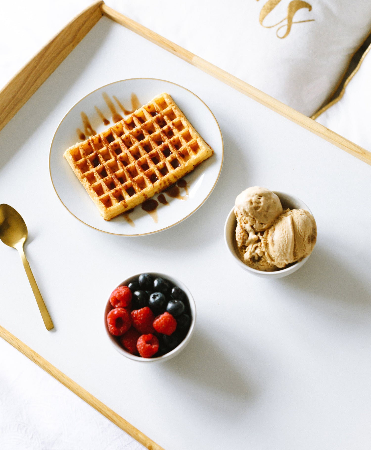 Waffel auf einem Teller, daneben eine Schüssel mit 3 Kugeln eis daneben eine Schale mit Himbeeren und Blaubeeren. Drapiert mit einem goldenen Löffel auf einem Tablett. Steht symbolisch für Fruchtzucker in industriellen Lebensmitteln. 