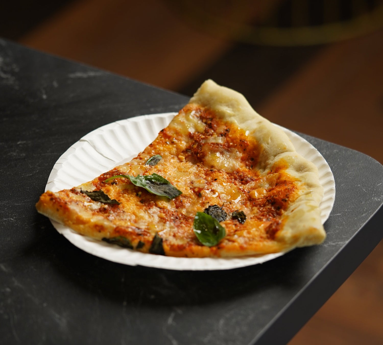 Stück Pizza auf einem Pappteller auf einem dunklen Steintisch steht symbolisch für industrielle Lebensmittel. 