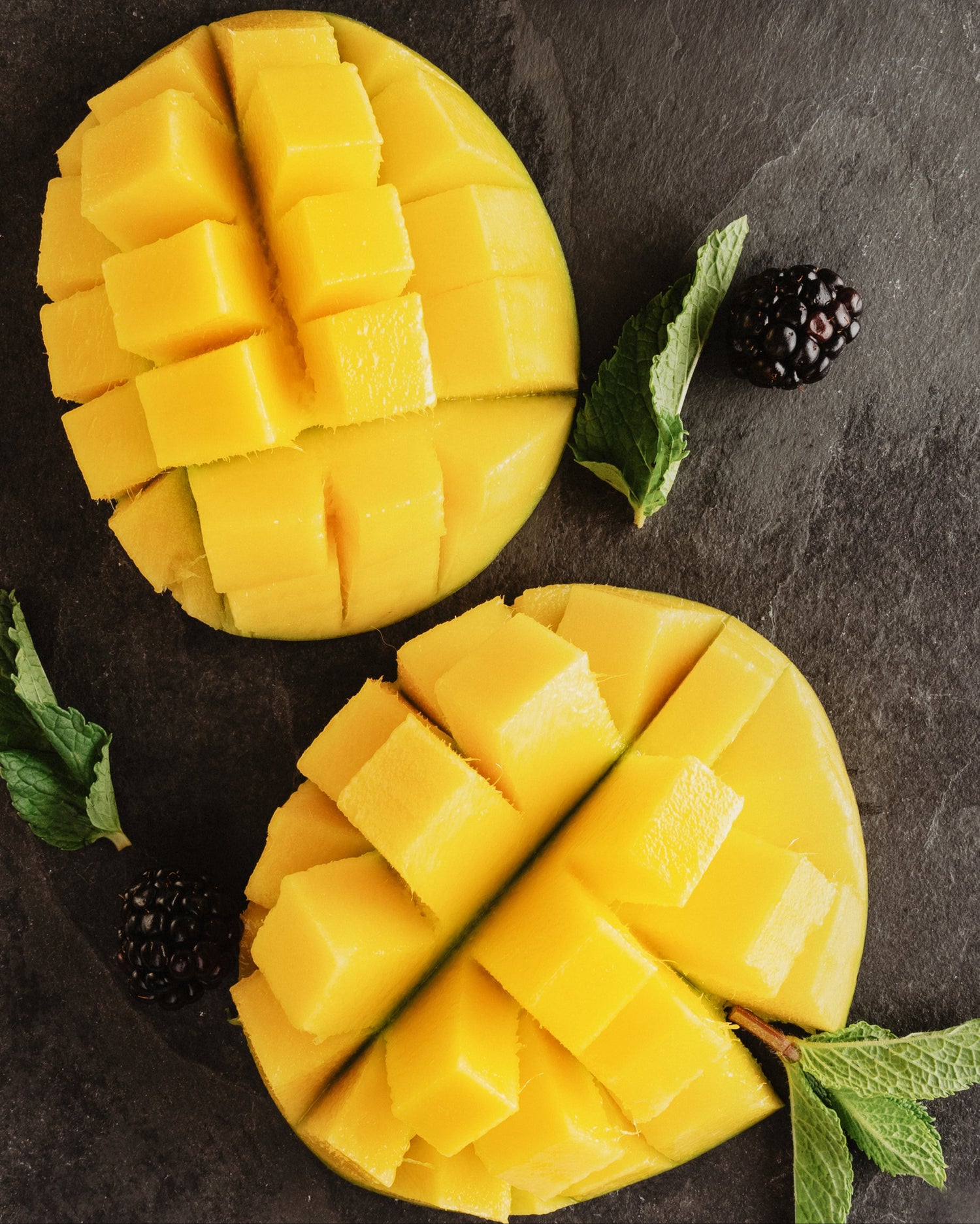 Zwei Mango Hälften in kleine quadratische Stücke geschnitten und mit Brombeeren und Blättern auf einer dunklen Holzunterlage drapiert. Steht symbolisch für Fruchzuckergehalt in Obstsorten